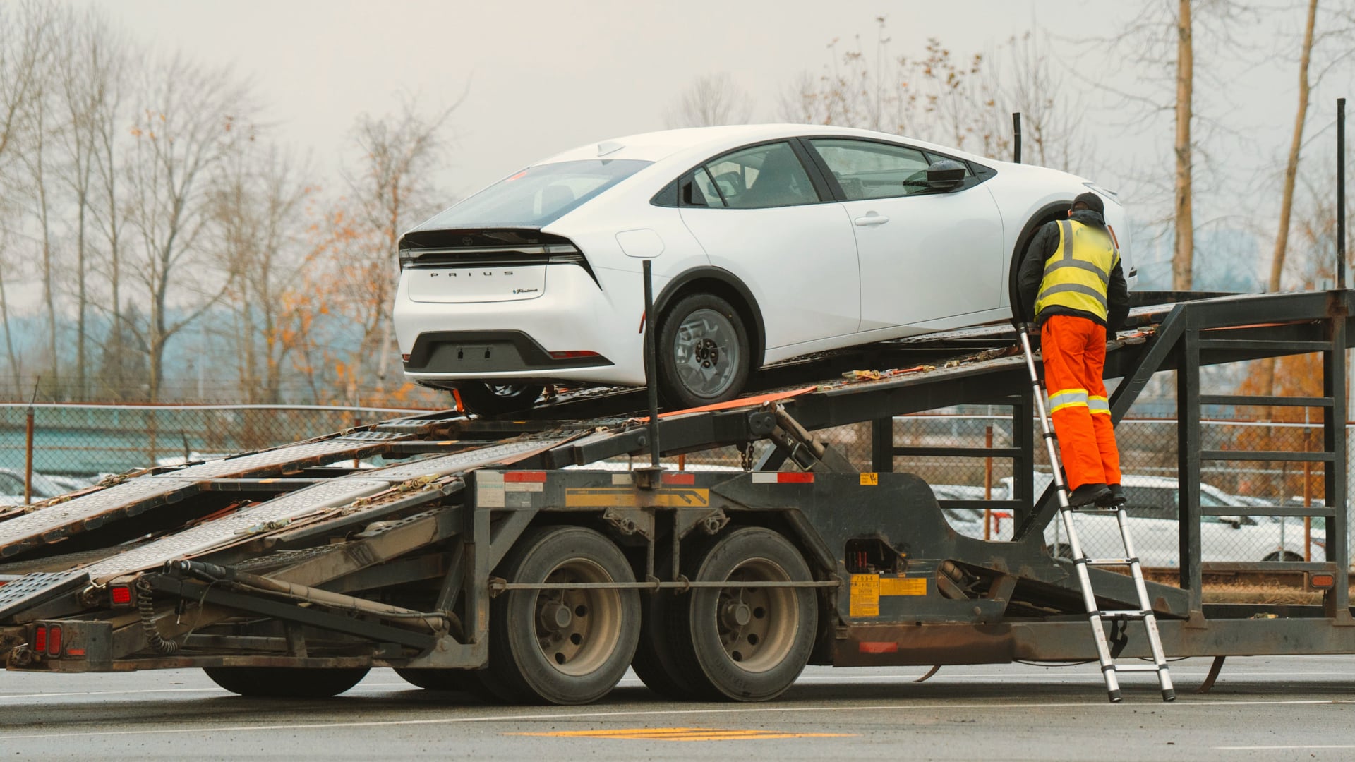 A worker in high-visibility clothing loading a white sedan onto an open car carrier on an overcast day