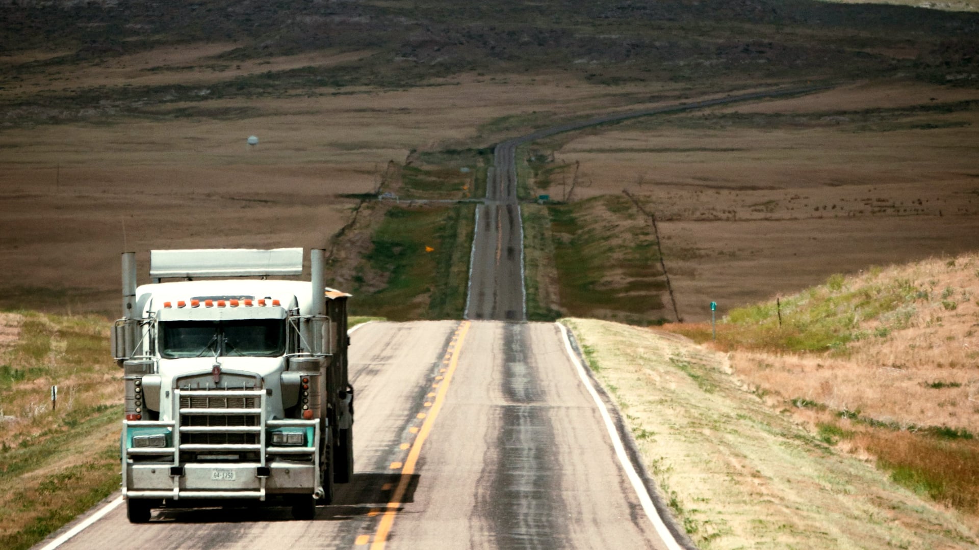 A semi-truck on a long, straight two-lane highway through open grassland